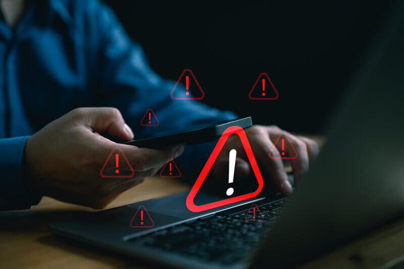 A business professional holding a smartphone while red warning symbols appear on a laptop screen, representing a ransomware attack delaying payroll and the financial risk to employees.